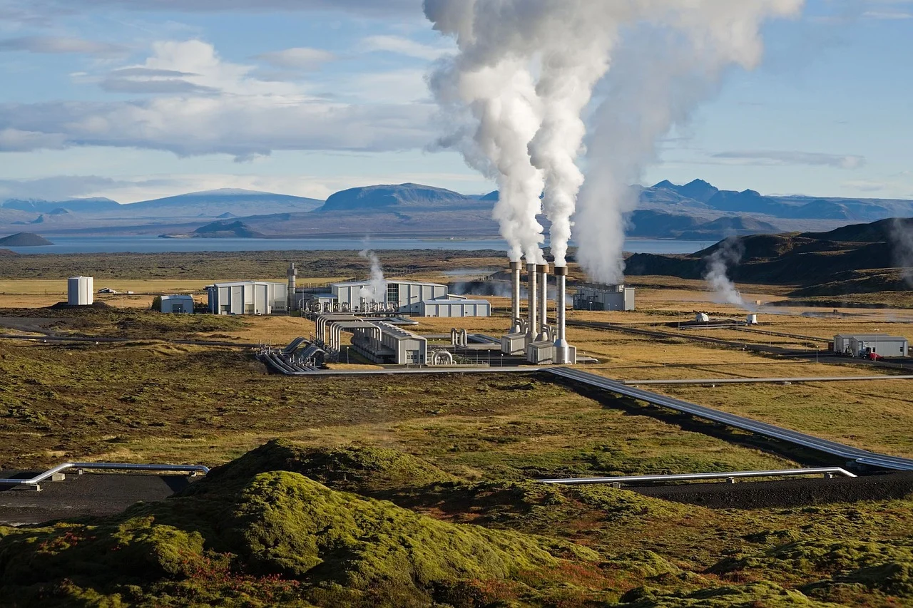 Geothermal energy plant in green and brown pasture with steam rising from stacks, mountains in the background